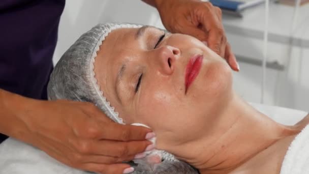 Seniorin lächelt, während Kosmetikerin ihr Gesicht mit Wattepads reinigt - Filmmaterial, Video