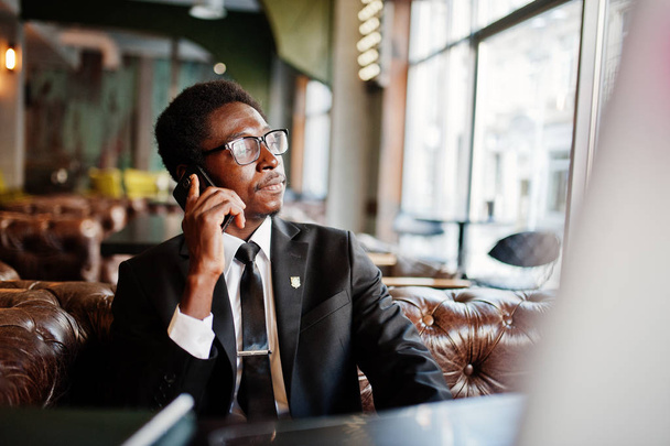 Business african american man wear on black suit and glasses sitting at office and speaking on phone behind laptop. - Foto, Imagem