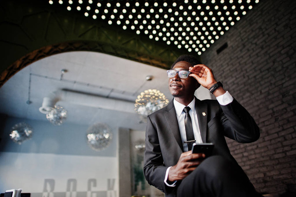 Business african american man wear on black suit and glasses at office looking on mobile phone against futuristic lights. - Fotó, kép