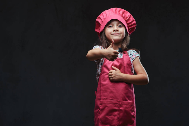 Cute little girl dressed in pink cook posing with thumbs up in a studio. Isolated on dark textured background. - Photo, image