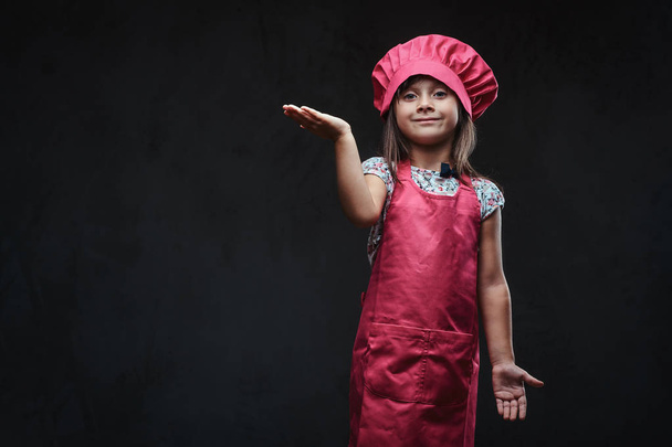 Happy little girl dressed in pink cook posing in a studio. Isolated on dark textured background. - Photo, image