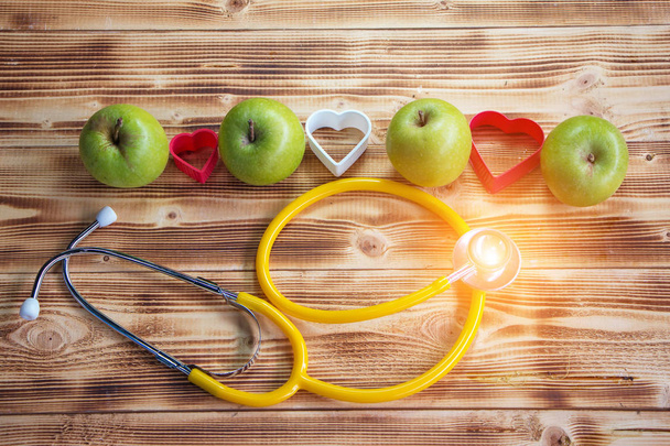 The green apple with heart plastic model stack on wooden timber board,at the back of yellow stethoscope,vintage warm light tone,blurry light around,health care concept - Photo, Image