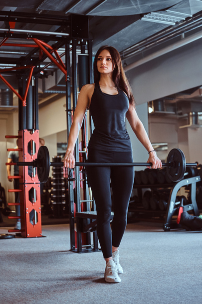 schöne brünette Frau in Sportbekleidung hält eine Langhantel während des Trainings im Fitnessclub oder Fitnessstudio. - Foto, Bild