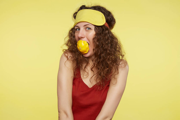 Young adult woman poses against yellow studio wall, promoting healthy lifestyle. Curly haired female starts morning from organic vegetables, holding scallop in mouth and wears sleep mask on head.  - Фото, изображение