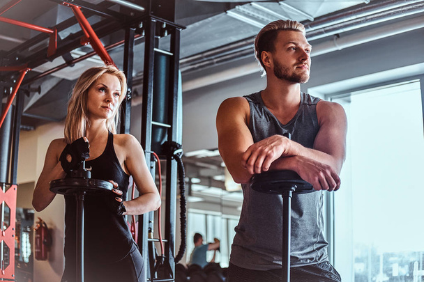 Portrait of an attractive sportive couple posing while leaning on barbells, looking away in the fitness club or gym. - Photo, Image