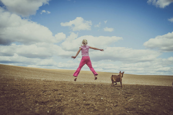 μικρό κορίτσι άλματα και σκύλος ζώο μαζί πεδίο στην ύπαιθρο κάτω από την γραφική συννεφιασμένο ουρανό ματ τόνο - Φωτογραφία, εικόνα