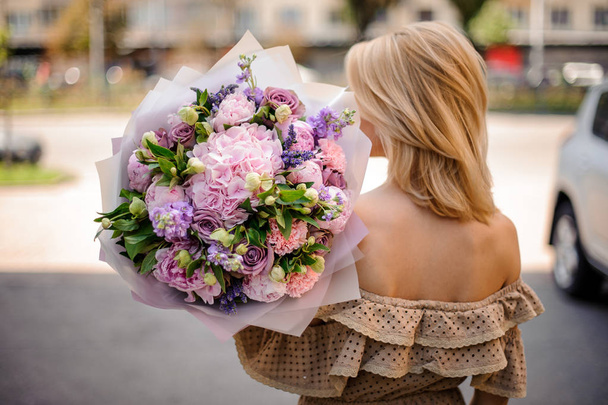 Rear view young beautiful girl in beige dress holding in her hands a bouquet of tender pink and violet flowers decorated with green leaves - Foto, immagini