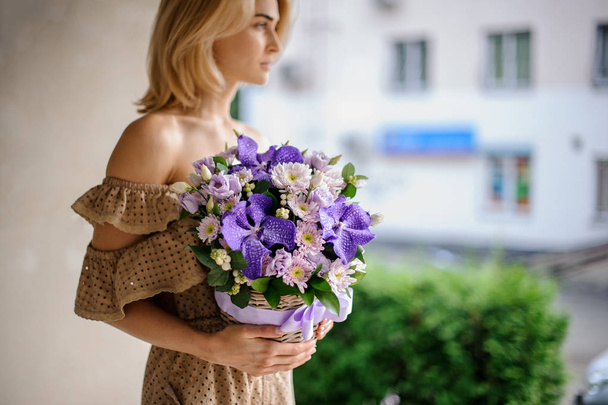 Young beautiful girl in beige dress holding in her hands a basket of pink chrisantemum and violet orchids decorated with green leaves - Foto, Imagen
