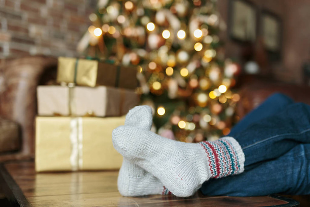 Adult person sitting on a sofa dressed Christmas socks. Golden lights on Christmas tree on the background. - 写真・画像