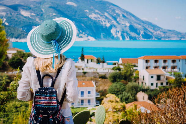 Tourist woman with blue sunhat and travel backpack admire view of colorful tranquil village Assos on sunny day. Visiting Kefalonia during summer time on Greece travel vacation - Zdjęcie, obraz