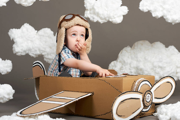 the boy plays in an airplane made of cardboard box and dreams of becoming a pilot, clouds from cotton wool on a gray background, retro style - Foto, Imagen