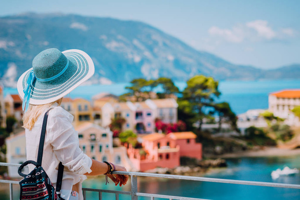 Tourist woman wearing blue sunhat and white clothes enjoying view of colorful tranquil village Assos on sunny day. Stylish female visiting Kefalonia in summer time on Greece travel vacation - Zdjęcie, obraz