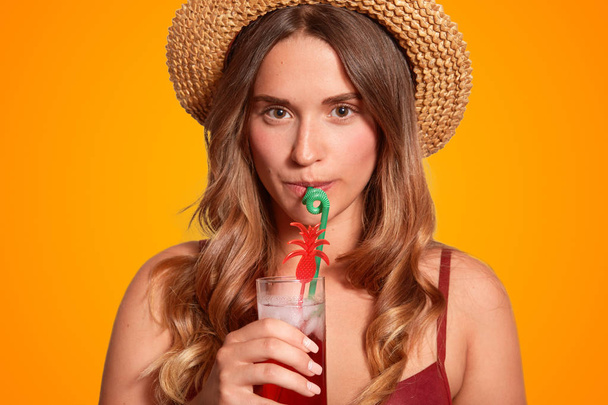 Cropped close up shot of serious pleasant looking female holds cold fresh beverage, drink with straw, has long hair, enjoys good summer rest, isolated over orange background. Recreation concept - Foto, Bild