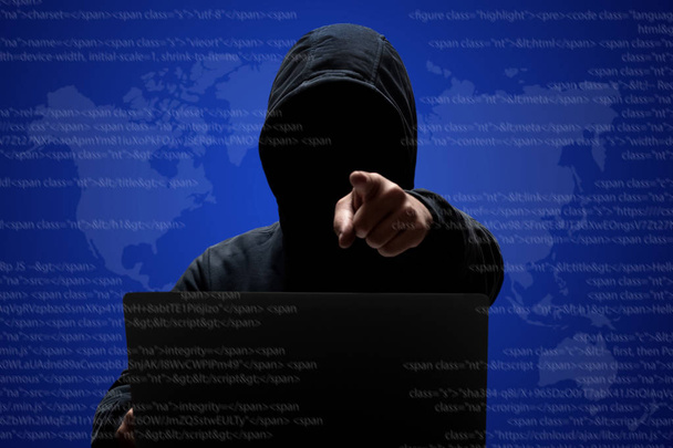 männlicher Hacker mit dunklem Gesicht in Kapuzenpulli, steht vor Laptop-Computer, zeigt auf Kamera, stiehlt Daten, lädt private Informationen herunter, posiert vor digitalem Interface-Hintergrund. Cyberspace. - Foto, Bild