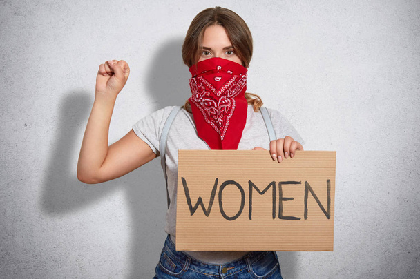 Feminism concept. Self confident young woman feminist protects womens rights, takes part in protest, keeps hands in fist raised, wears red bandana and grey t shirt, stands indoor, being serious - Foto, imagen