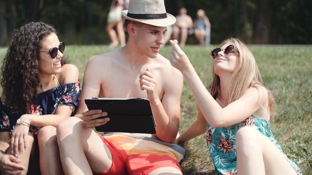 Friends looking on their tablet while sitting on a grass. Happy mixed ethnicity group of friends using computer tablet, outdoors. - Foto, Imagen