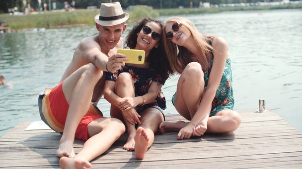 Group of friends making selfies by the lake. Cheerful friends spending time by the lake and using phone. - Zdjęcie, obraz