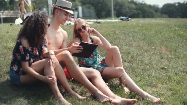 Friends looking on their tablet while sitting on a grass. Happy mixed ethnicity group of friends using computer tablet, outdoors. - Video, Çekim