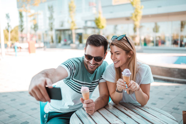 Porträt eines jungen Paares, das ein Selfie macht, während es draußen sitzt und Eis in einer Tüte isst - Foto, Bild