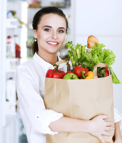 Sebzeli alışveriş torbası tutan genç bir kadın. Mutfakta duruyor. Mutfakta kameraya bakan bir kadın var. - Fotoğraf, Görsel
