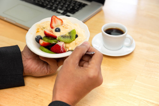 Businessman having bowl of fresh fruits salad with yogurt and coffee and laptop on table. - Photo, Image