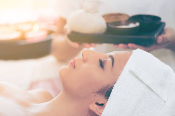 Relaxed woman lying on spa bed prepared for spa massage with therapist holding spa treatment set in background. Luxury wellness, stress relief and rejuvenation concept. - 写真・画像