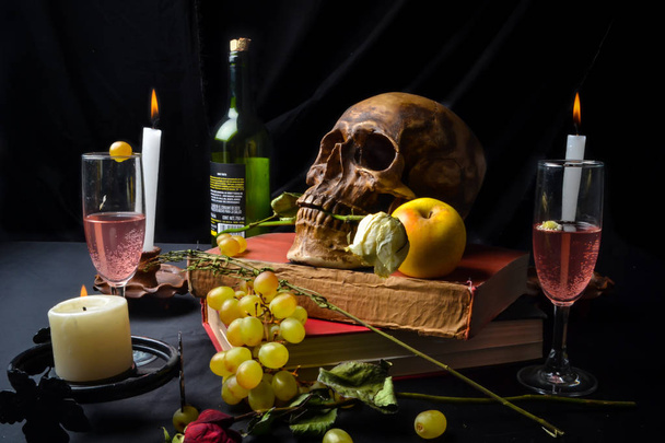 Naturaleza muerta con una calavera con rosas, libros, frutas, vino y velas sobre un fondo oscuro - Foto, afbeelding