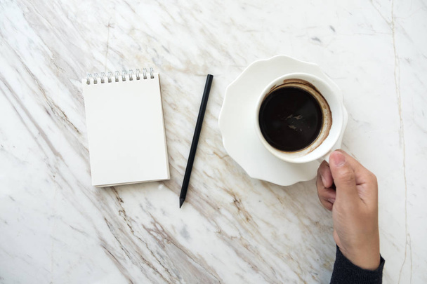 Widok z góry obraz dłoni trzymającej filiżankę kawy z pustym białym notebookiem na stole - Zdjęcie, obraz