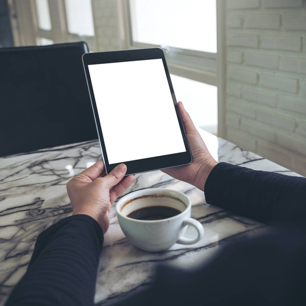 Изображение рук, держащих черный планшет с чистым белым экраном и чашкой кофе на столе в кафе  - Фото, изображение