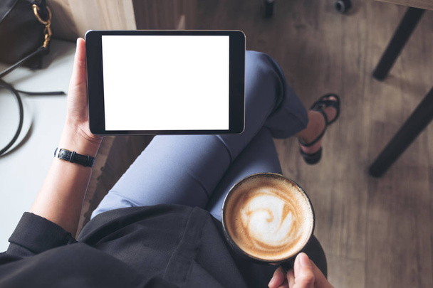 Imagem Mockup de uma mulher sentada de pernas cruzadas e segurando pc tablet preto com tela de desktop branca em branco enquanto bebe café no café - Foto, Imagem