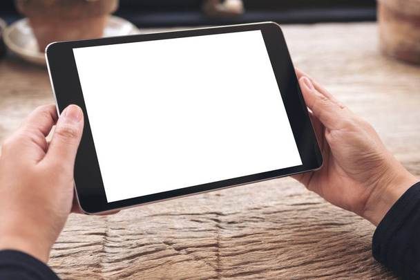 Imagen burlona de las manos sosteniendo la tableta negra PC con pantalla blanca en blanco en la mesa de madera vintage  - Foto, Imagen