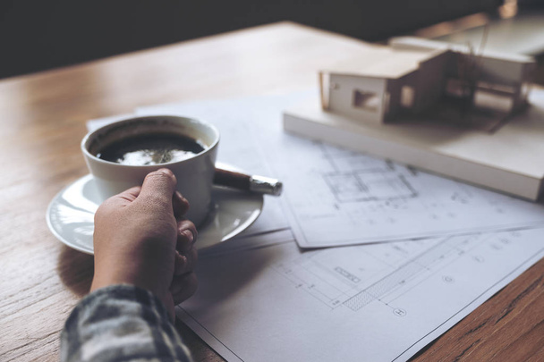 Un arquitecto trabajando en un modelo de arquitectura con papel de dibujo de tienda mientras bebe café en la oficina - Foto, Imagen