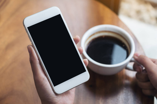 Mockup-Bild der Hände einer Frau, die ein weißes Handy mit leerem schwarzen Bildschirm hält, während sie im Café Kaffee trinkt - Foto, Bild