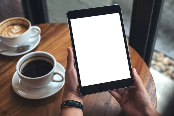 Imagen burlona de manos sosteniendo PC tableta negra con pantalla blanca en blanco y tazas de café en la mesa  - Foto, Imagen