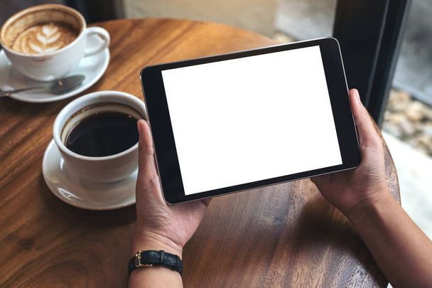 Макетне зображення рук, що тримають чорний планшетний ПК з білим порожнім екраном і чашками кави на столі
  - Фото, зображення