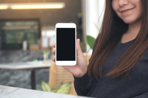 Imagen burlona de una mujer sosteniendo y mostrando un teléfono móvil blanco con pantalla negra en blanco en la cafetería - Foto, Imagen