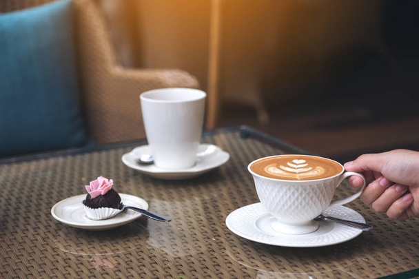 Closeup image of a hand holding a cup of hot latte coffee with latte art and snack on table in cafe - Fotó, kép