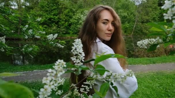 鳥桜の開花のカメラにポーズかなりヨーロッパの女の子モデル。長いふわふわの髪と彼女の頭の上の枝を持つ若い女性は公園、スローモーションで撮影します。 - 映像、動画