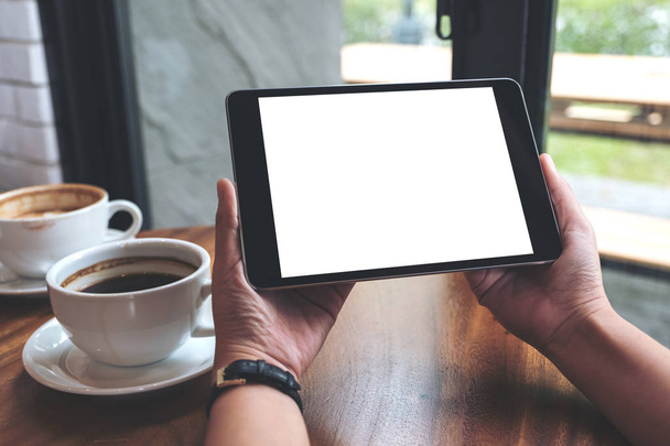 Mockup-Bild der Hände hält schwarzen Tablet-PC mit weißen leeren Bildschirm und Kaffeetassen auf dem Tisch  - Foto, Bild