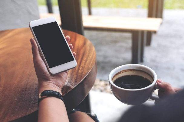 Mockup image of hands holding white mobile phone with blank black screen while drinking coffee in cafe - Foto, afbeelding