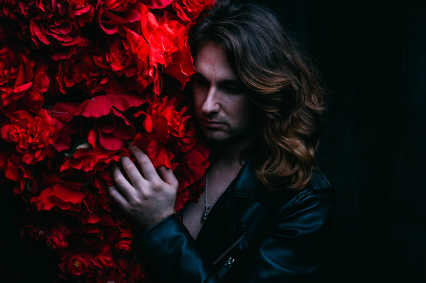 brutal man in a leather jacket on a naked body with long hair stands near the decor with red flowers flowers. a lonely man is sad and desperate - Foto, afbeelding