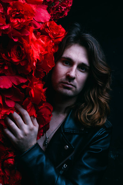 brutal man in a leather jacket on a naked body with long hair stands near the decor with red flowers flowers. a lonely man is sad and desperate - Fotó, kép