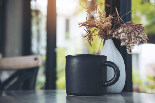 Una taza negra de café caliente con flores secas en un jarrón blanco sobre la mesa en la cafetería moderna
 - Foto, imagen