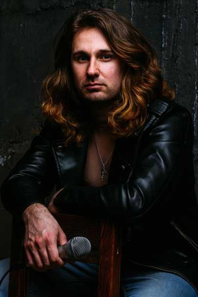 a portrait of a brutal, handsome man, a singer who sits on a chair on a dark background, in a black leather kurt on his naked body and holds a microphone in his hands. presenter, singer at the concert - Foto, Imagen