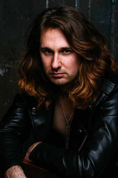 a portrait of a brutal, handsome man, a singer who sits on a chair on a dark background, in a black leather kurt on his naked body and holds a microphone in his hands. presenter, singer at the concert - Фото, зображення