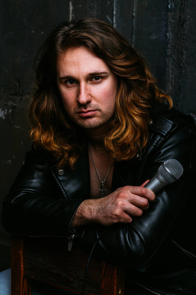 a portrait of a brutal, handsome man, a singer who sits on a chair on a dark background, in a black leather kurt on his naked body and holds a microphone in his hands. presenter, singer at the concert - Foto, Imagen
