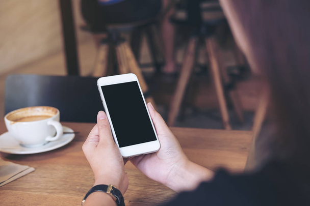 Mockup image of hands holding white mobile phone with blank black screen with coffee cups on wooden table in restaurant - Photo, Image