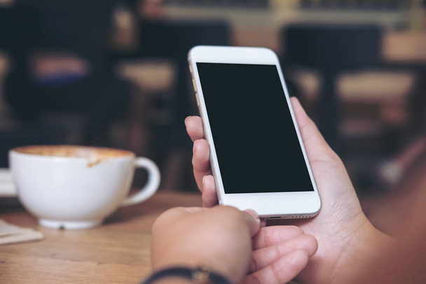 Изображение рук, держащих белый мобильный телефон с чистым черным экраном с чашками кофе на деревянном столе в ресторане
 - Фото, изображение
