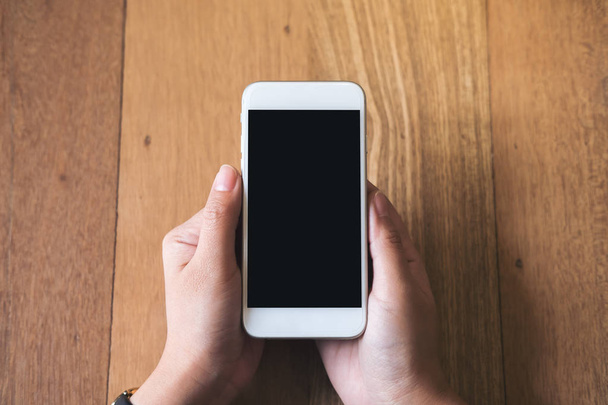 Imagem Mockup das mãos da mulher segurando telefone celular branco com tela preta em branco no fundo da mesa de madeira vintage
 - Foto, Imagem