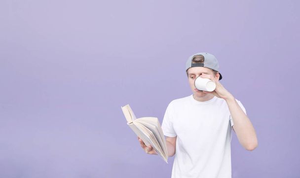 Portrait of a young man in a cap and a white T-shirt who reads a book and drinks coffee on a pastel purple background. Student reads a book and drinks coffee on the background of a purple wall. - Valokuva, kuva